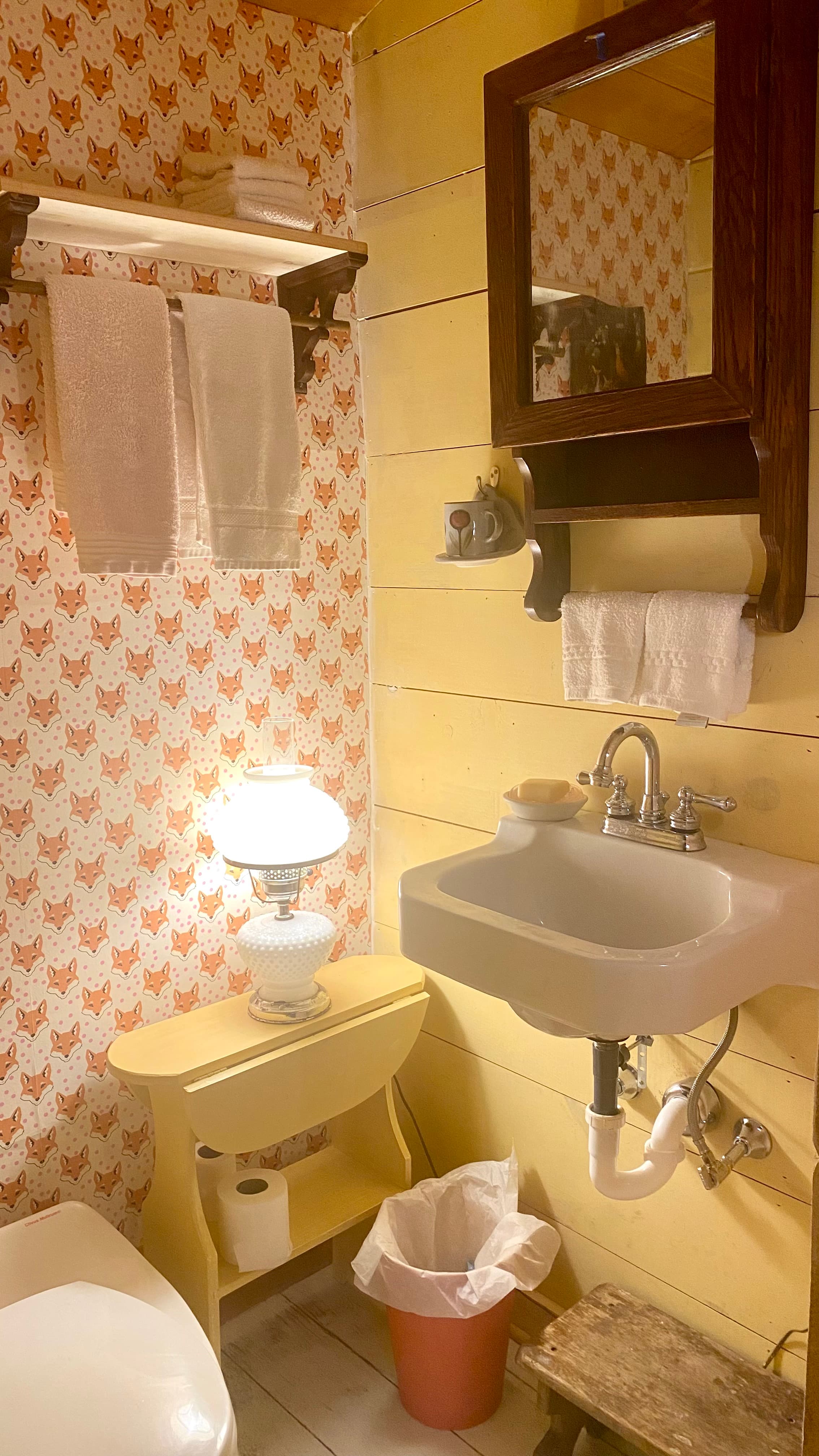 small bathroom with a white Clivus Multrum toilet, a wall hung sink and a yellow drop leaf table with a milk glass lamp. The wall paper on 2 walls has orange and black fox faces on it's white background