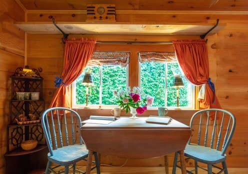Maple kitchen table with 2 blue wooden chairs set before a double window with brick colored curtains and a blue and brick colored set of cloth shades
