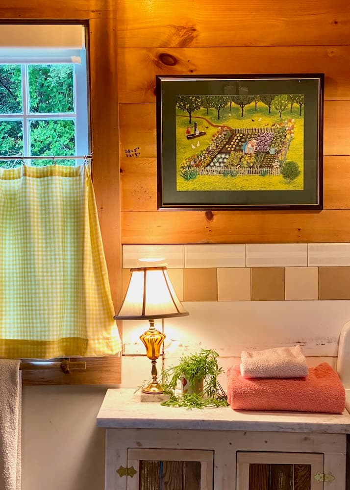 A cozy bathroom featuring a window with a yellow checkered curtain, a framed garden painting, a lamp, and neatly stacked towels.