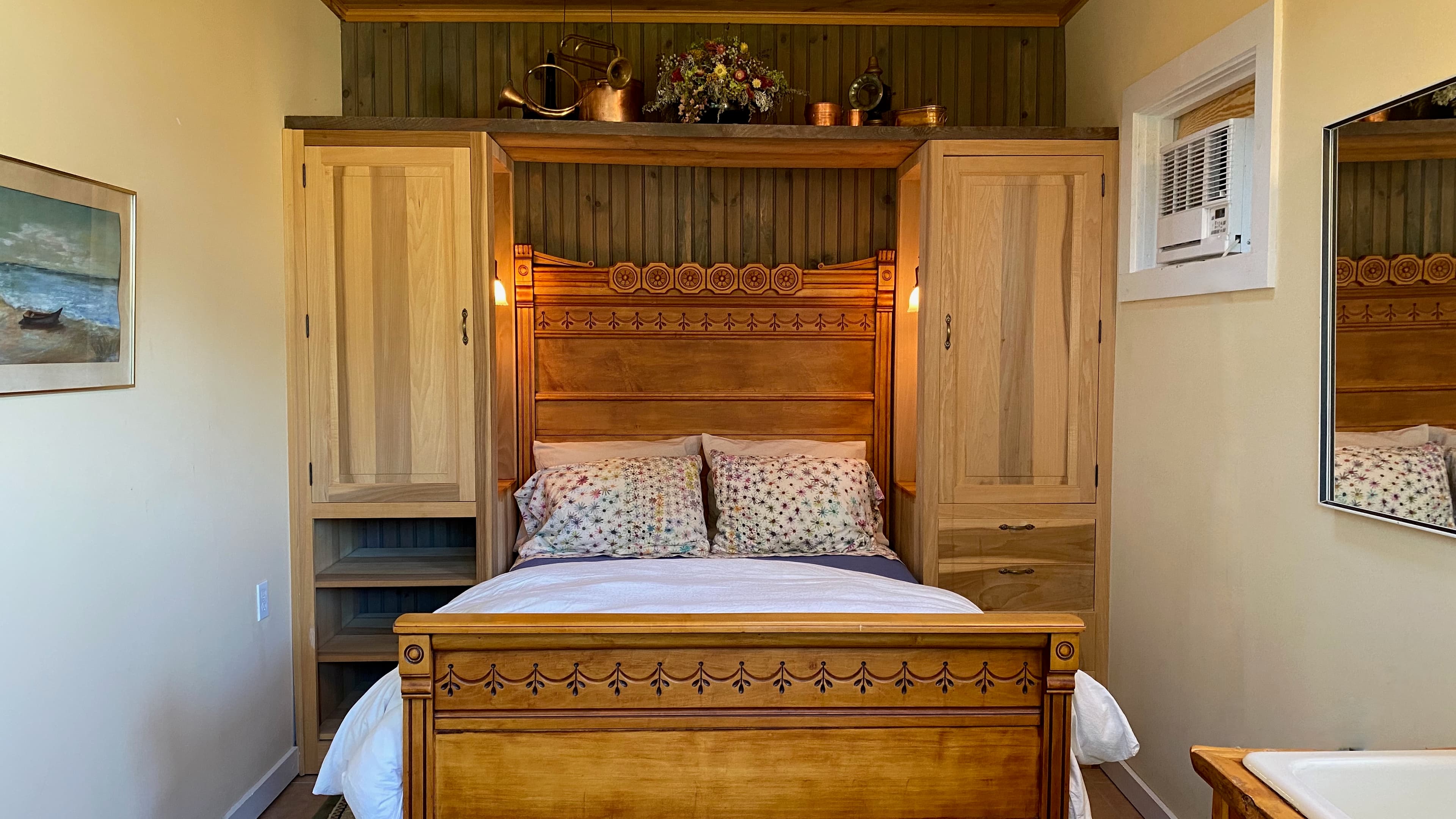 Double bed tucked between 2 floor to almost ceiling cabinets. Bed has antique carved headboard and footboard with a natural honey colored finish. Room has pale yellow walls and cabinetry is unfinished wood. Decorative brass objects on display in the shelf above.