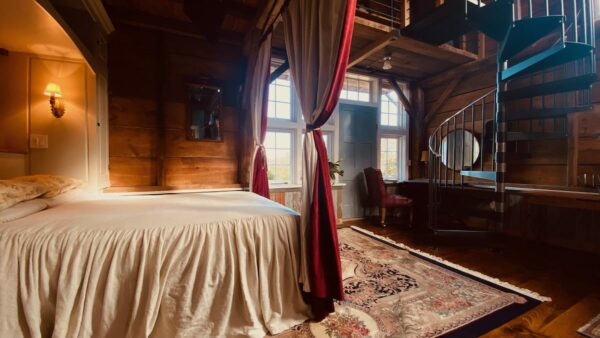 and a Queen sized bed with a beige linen bedspread.|View of an antique carriage house barn living room with a brick fireplace centered in the room a comfy couch