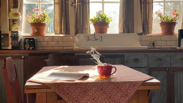 Carriage House kitchen table set with red handwoven tablecloth and a steaming cup of tea and newspaper on tabletop. 3 windows ihn the background with geraniums blooming in terracotta pots.