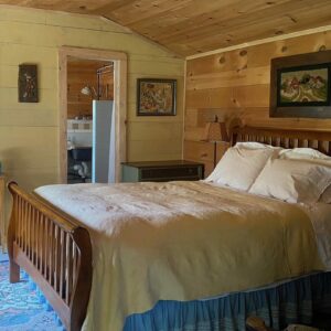 Queen size bed neatly made with yellow quilt and four pillows in the bedroom of the Garden Cottage. The room is sided in horizontal unpainted pine siding with assorted artwork and a large mirror. There is a large blue patterned rug on the floor.|Looking down at a bathroom with a beige and white checked floor
