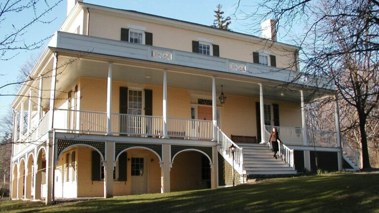 Exterior view of the Thomas Cole House. A 3 story beautiful yellow house with a large stairway and wrap around porch.